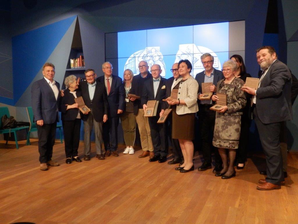 Zdjęcie przedstawia grupę odznaczonych medalem. Osoby z medalami w rękach stoją na tle ekranów ledowych w jednej z sal Muzeum Polin w Warszawie.